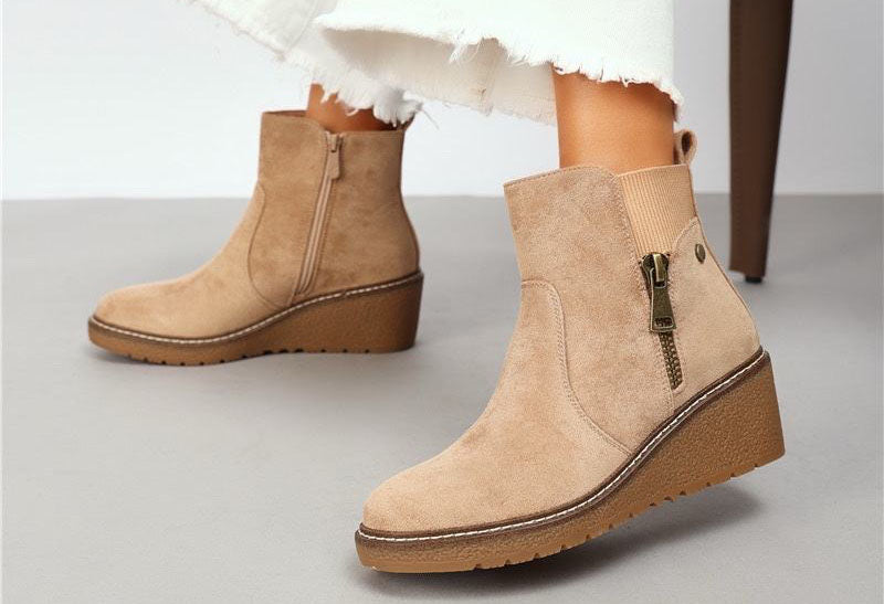 Beige faux suede wedge boot with decorative brass zip on a models foot
