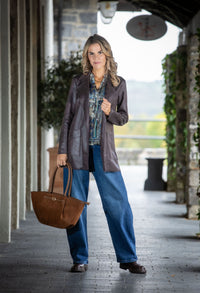Woman standing on a wooden deck holding a brown bag, wearing a dark jacket and blue jeans.