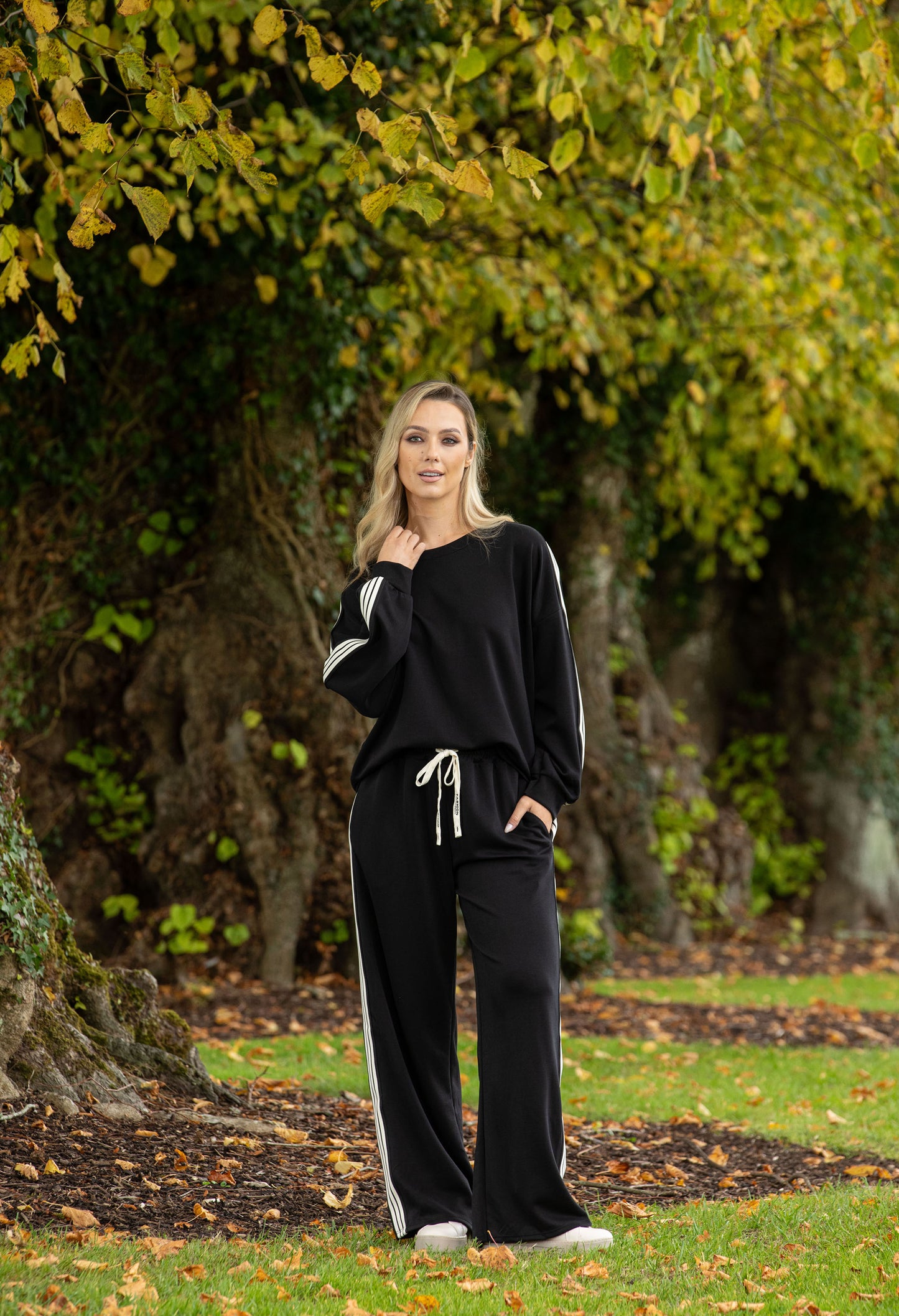 Woman in black outfit with white stripes standing in a park with trees and grass.