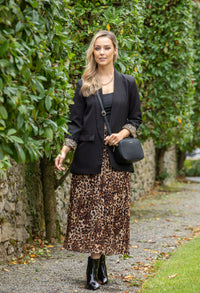 Woman in a black blazer and leopard print skirt standing outdoors with greenery in the background