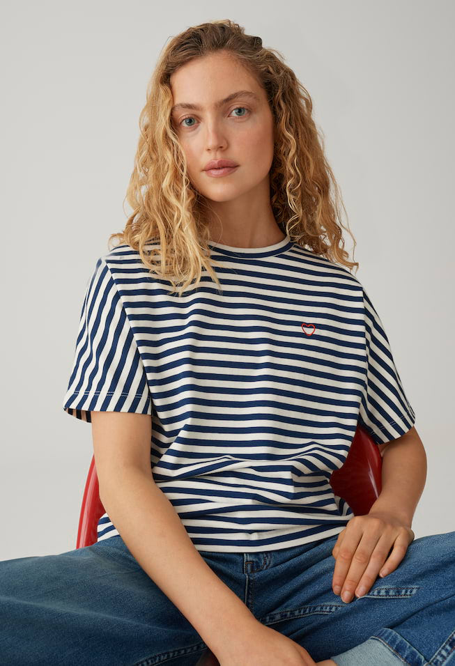Woman wearing a striped shirt with a small logo, sitting on a red chair against a plain background