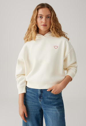 Person wearing a cream hoodie with a heart design on a plain background