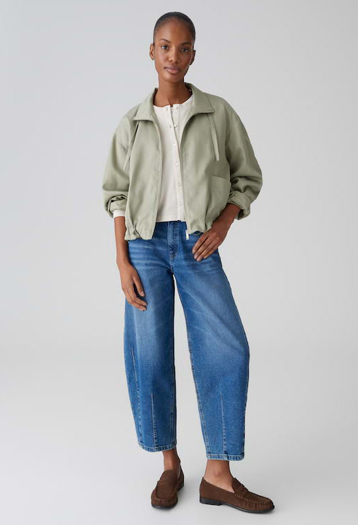 Woman wearing a light green jacket, white shirt, blue jeans, and brown shoes on a plain background