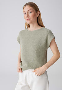 Woman wearing a green knitted top and white pants against a light gray background