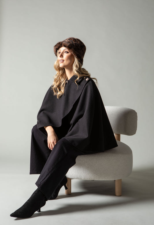 Woman wearing a black cape and hat sitting on a white chair against a gray background