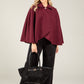 Woman wearing a burgundy cape and black pants holding a black handbag on a light gray background