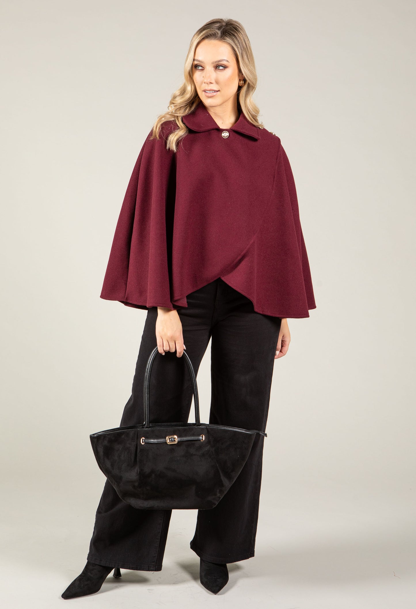 Woman wearing a burgundy cape and black pants holding a black handbag on a light gray background