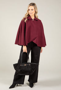 Woman wearing a burgundy cape and black pants holding a black handbag on a light gray background