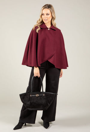 Woman wearing a burgundy cape and black pants holding a black handbag on a light gray background