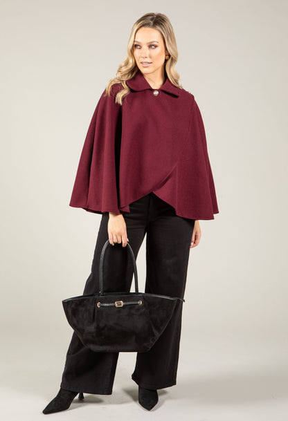Woman wearing a burgundy cape and black pants holding a black handbag on a light gray background