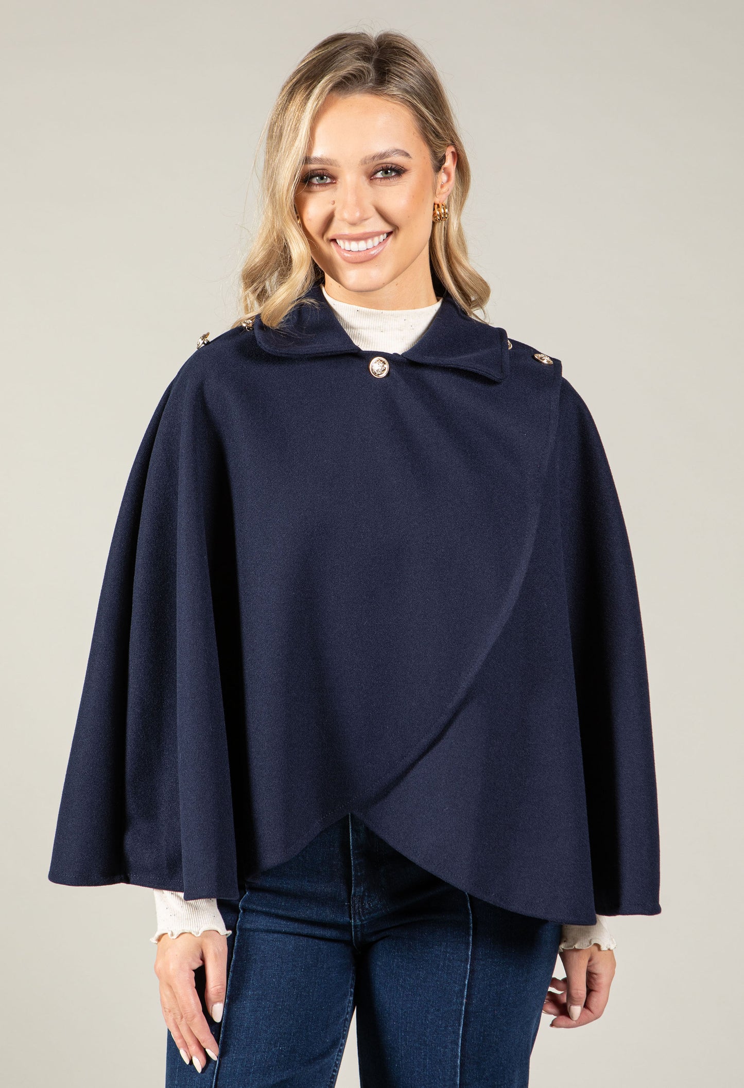 Woman wearing a navy blue cape over a white shirt and jeans on a plain background