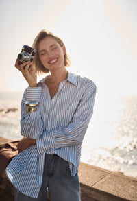 Woman holding a camera by a waterfront wearing a striped blue shirt