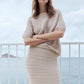 Woman wearing a beige knit dress standing by a railing with a blurred coastal background