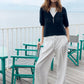 Woman standing on a wooden deck with teal chairs and a white railing, wearing a navy sweater and white pants.