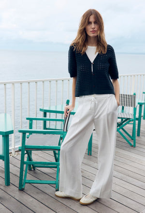 Woman standing on a wooden deck with teal chairs and a white railing, wearing a navy sweater and white pants.