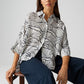 Woman sitting on a stool wearing a black and white patterned shirt and navy pants against a light gray background