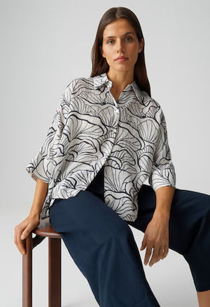 Woman sitting on a stool wearing a black and white patterned shirt and navy pants against a light gray background