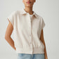 Woman wearing a beige sleeveless sweater over a white shirt with light blue jeans on a plain background