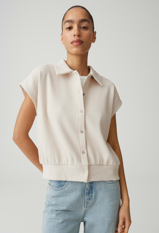 Woman wearing a beige sleeveless sweater over a white shirt with light blue jeans on a plain background