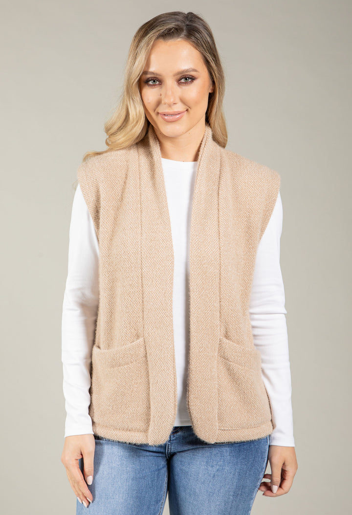 Woman wearing a beige vest over a white long-sleeve shirt and blue jeans on a beige background