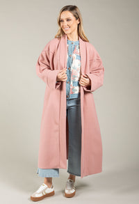 Woman wearing a long pink coat over a floral top and jeans on a plain background
