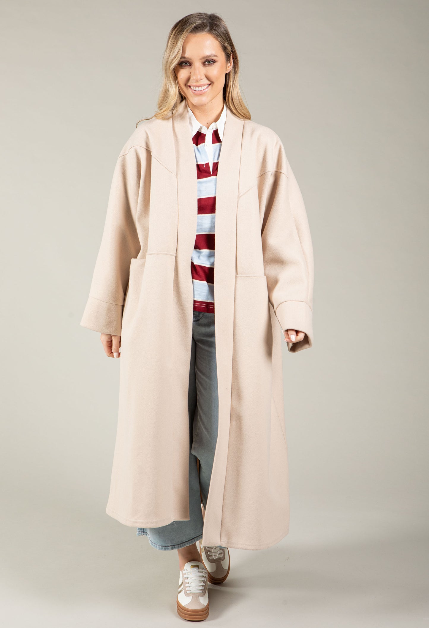 Woman wearing a long beige coat over a striped sweater on a plain background