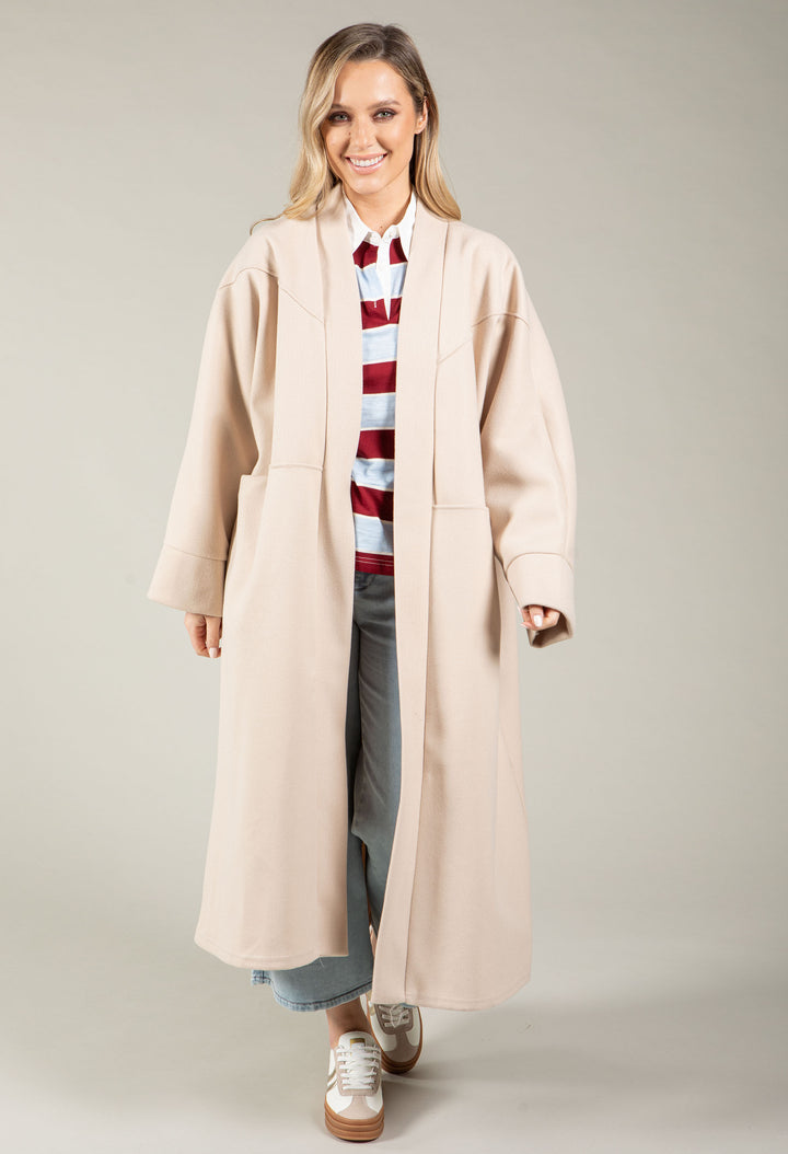 Woman wearing a long beige coat over a striped sweater on a plain background
