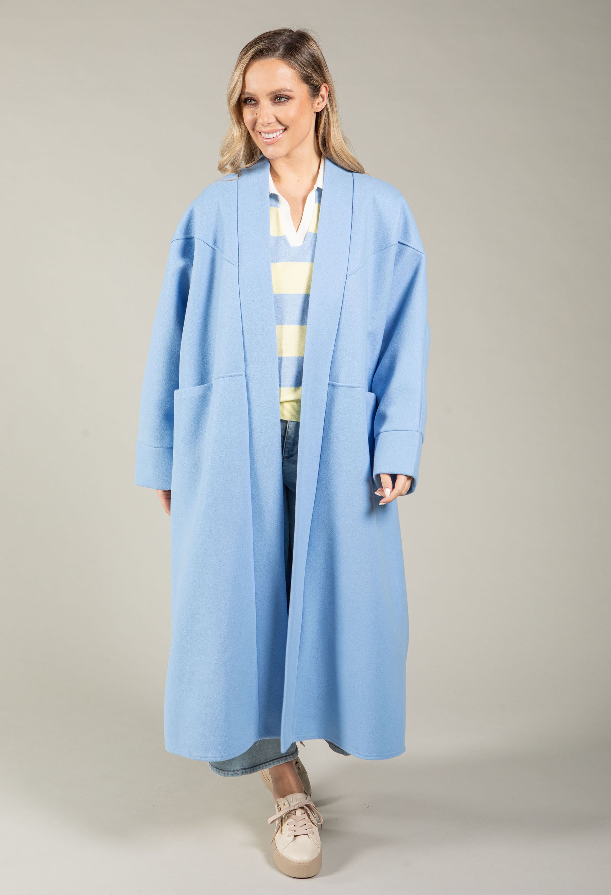 Woman wearing a long light blue coat over a striped shirt on a plain background