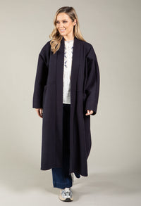 Woman wearing a long navy coat over a white top with a floral pattern, standing against a plain background.