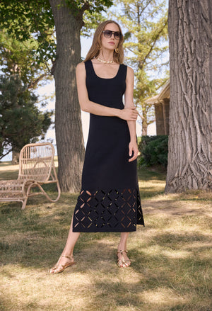 Woman in a black dress standing outdoors with trees and a chair in the background