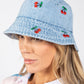 Woman wearing a light blue denim bucket hat with cherry embroidery on a white background