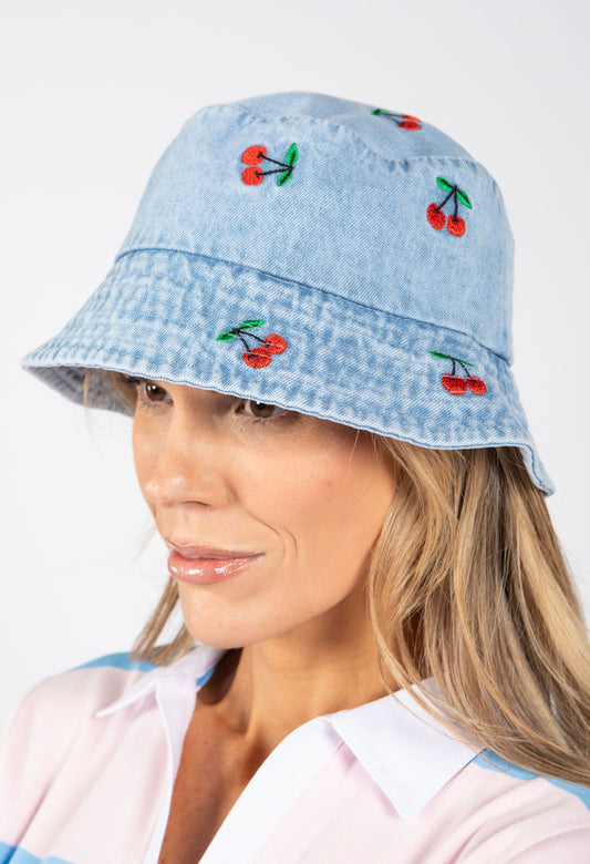 Woman wearing a light blue denim bucket hat with cherry embroidery on a white background
