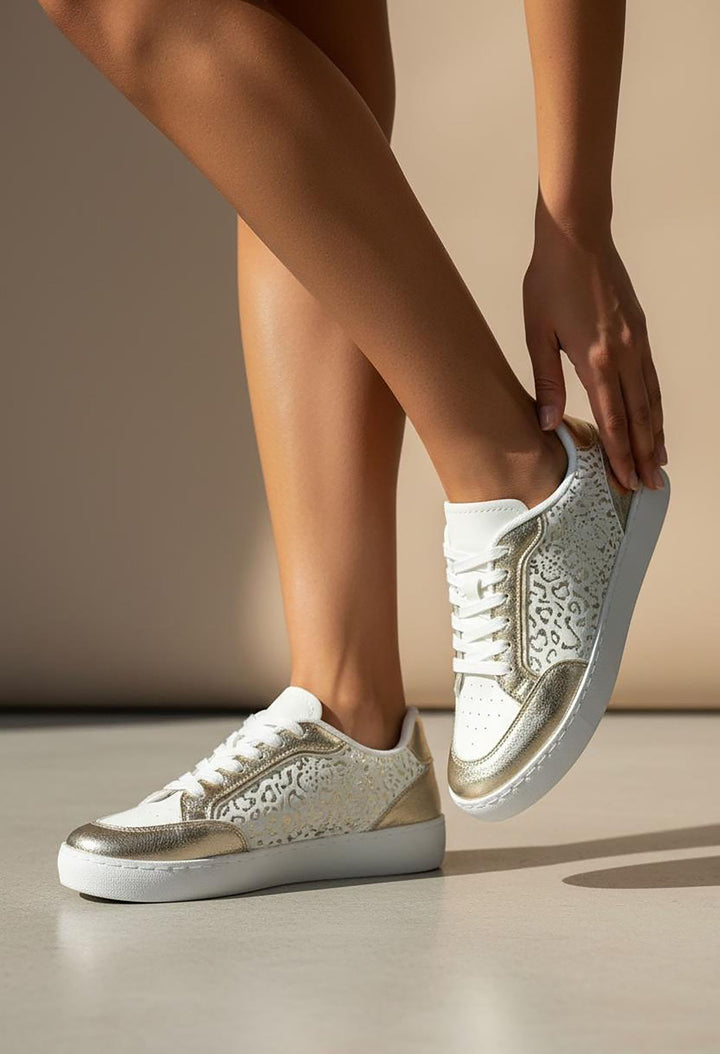 White sneakers with gold lace details worn by a person on a neutral background