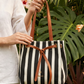 Person holding a black and white striped bag with brown leather straps against a green leafy background