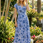 Woman in a blue floral dress standing in a garden with palm trees and flowers.