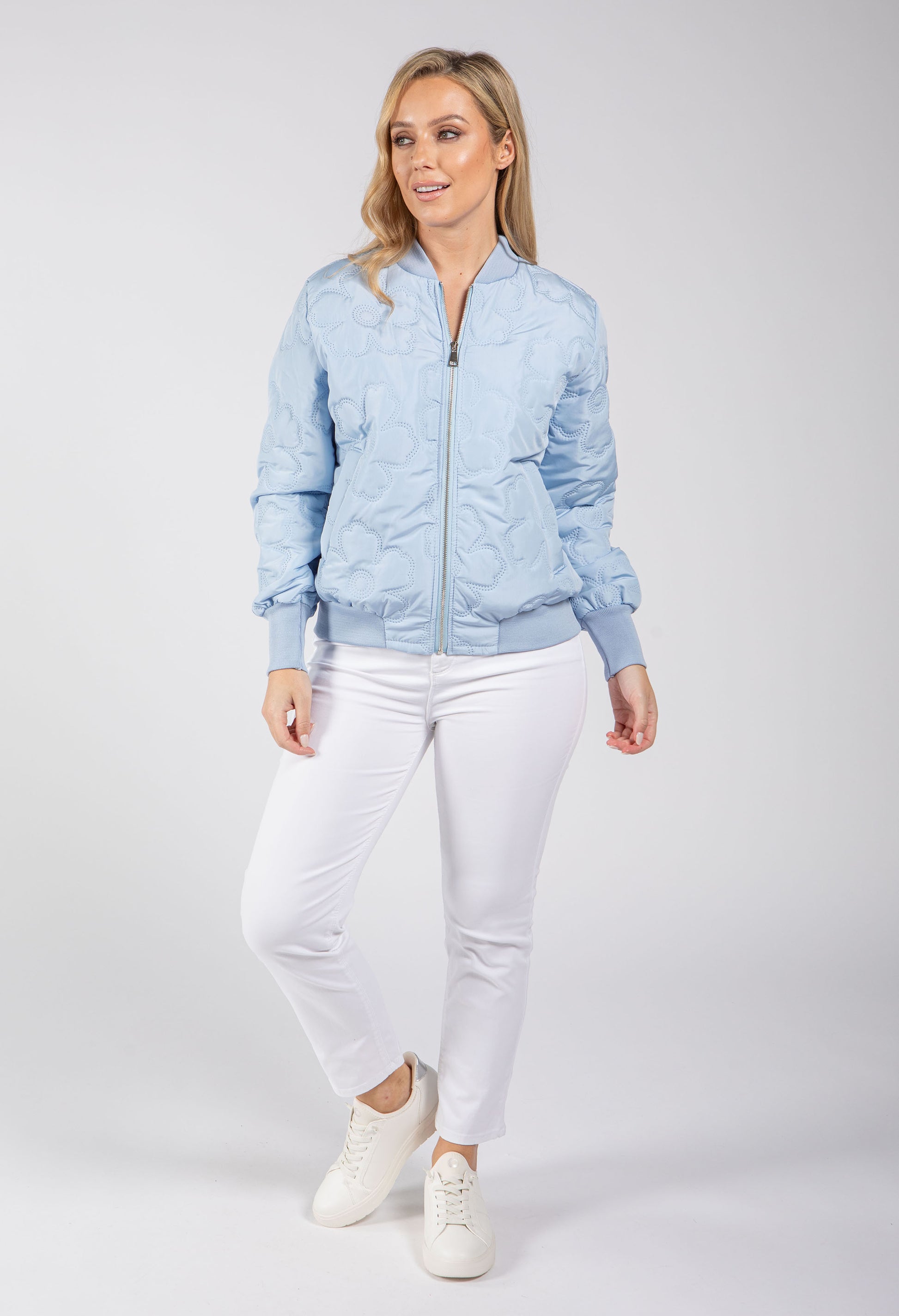 Woman wearing a light blue bomber jacket and white pants on a gray background