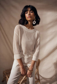 Woman wearing a beige knitted sweater and pants sitting on a chair against a plain background