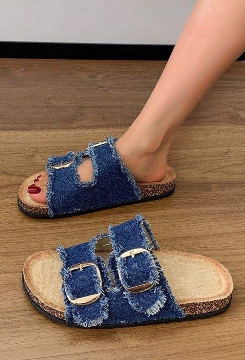 Denim sandals with buckles worn by a person on a wooden floor.