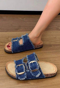 Denim sandals with buckles worn by a person on a wooden floor.
