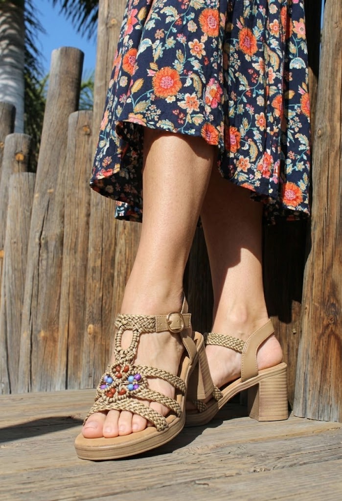 Person wearing a floral dress and beige sandals with embellishments on a wooden deck.