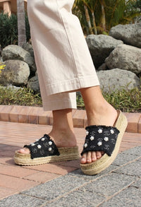 Person wearing black sandals with floral patterns on a stone path with rocks and plants in the background