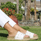 Person wearing white sandals with jute sole on grass with palm trees and flowers in the background