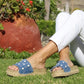 Person wearing blue sandals with white patterns on grass with a red pot and plants in the background