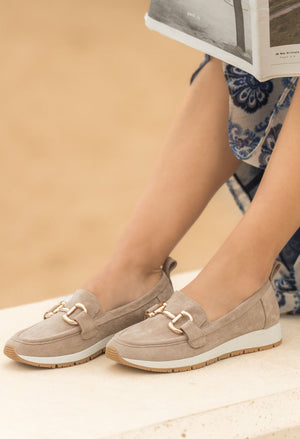 Beige loafers worn by a person sitting on a wooden floor with a beige background