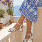Person wearing a blue and white patterned dress and white sandals on a balcony with ocean view.