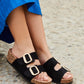 Black sandals with cork soles worn by a person in a blue dress on a stone surface.