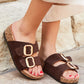 Brown sandals with gold buckles worn by a person on a stone pavement.