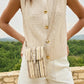 Person wearing a beige knitted vest over a white shirt with gold buttons, holding a straw handbag, outdoors.