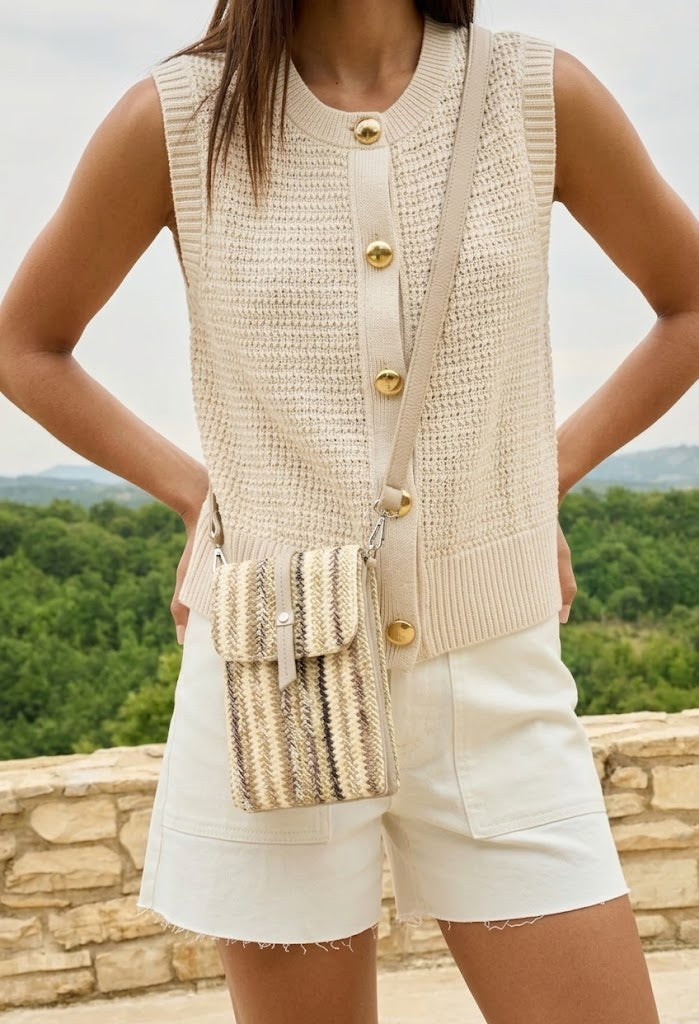 Person wearing a beige knitted vest over a white shirt with gold buttons, holding a straw handbag, outdoors.