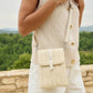Person wearing a beige sleeveless top with gold buttons and holding a matching woven bag, standing against a scenic background.
